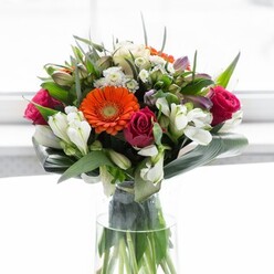 Brimming with character and crafted with artistry, this bouquet embodies both the urban sophistication and historic richness found across Aldgate. Vibrant orange gerberas and blushing peach counterparts anchor the design, their gently rounded petals encircling prominent brown centers and summoning the lively energy of Petticoat Lane Market. Lush fuchsia roses, each tightly wound, add a sense of passion and depth, while elegant sprays of white alstroemeria and petite spray roses introduce a fresh, airy texture, suggesting the gentle calm found in small green oases hidden amid Aldgate's cityscape. Subtle notes of soft pink and clusters of tiny, airy white blossoms bring depth and variety, the arrangement filled out with a dynamic selection of glossy and pointed greenery. Broad aspidistra leaves, elegantly curved, form a nurturing green base as steady as the foundations of Aldgate's historic streets, further enhancing the full, rounded silhouette. The tall, slender glass vase allows the vibrant stems to shine, reinforcing the sense of freshness and contemporary style. Against a softly lit white background, every bloom and leaf is celebrated, offering an invitation to joy, gratitude, and connection. This arrangement is perfectly suited for events near Fenchurch Street or thoughtful deliveries across Aldgate, its every element a testament to the care and professionalism of local expert florists.